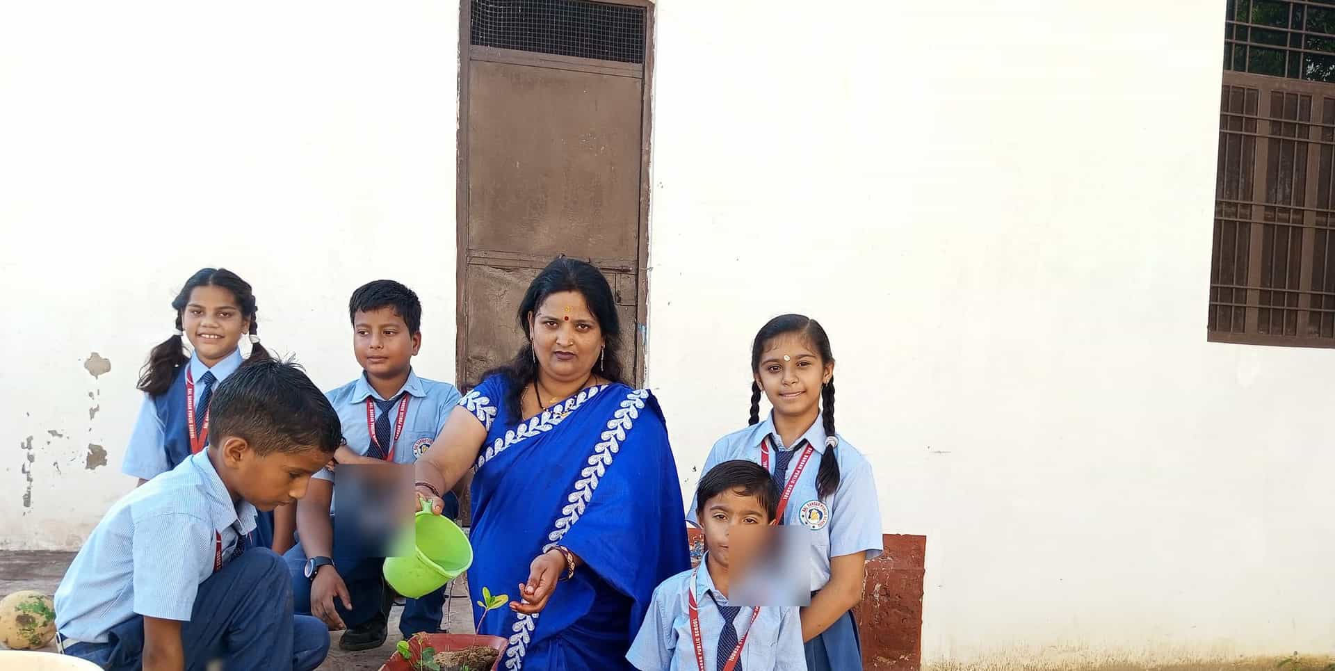 A woman waters a sapling in a red pot while five children in school uniform gather round her outside the school