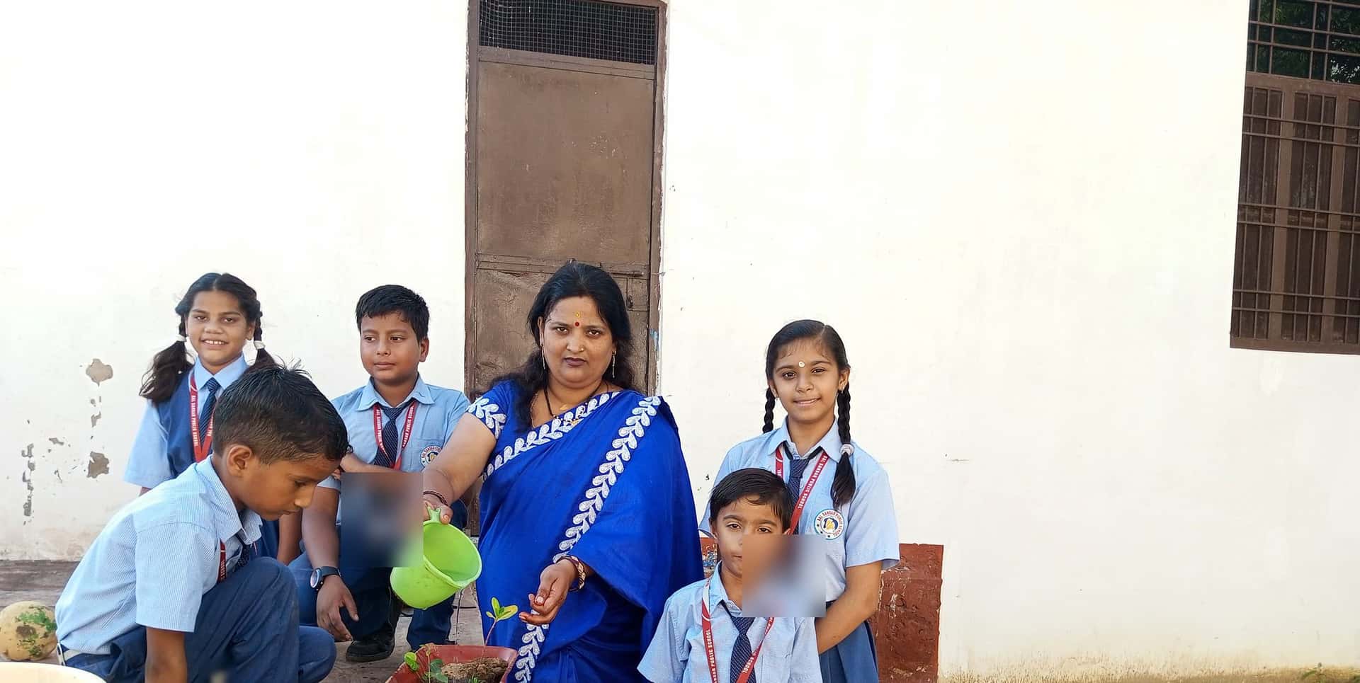 A woman waters a sapling in a red pot while five children in school uniform gather round her outside the school