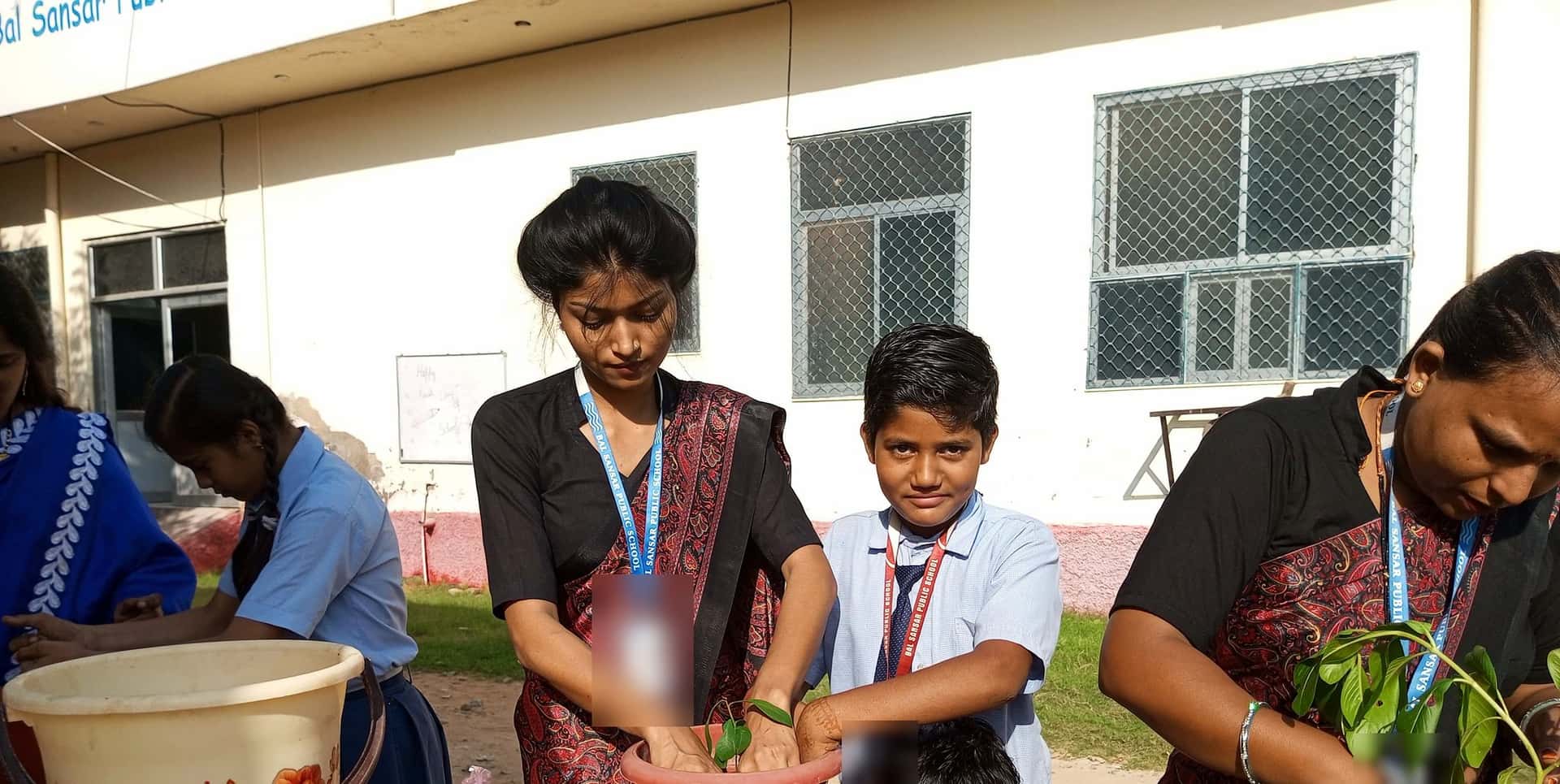 A woman and a student press soil around a sapling in a red pot as a younger boy looks on