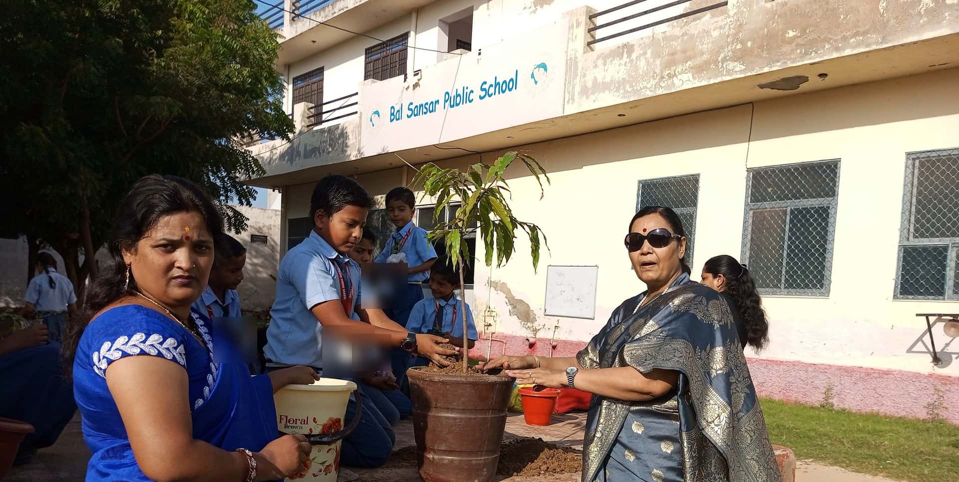 Students and two women plant a tall sapling in a large pot beneath the Bal Sansar Public School sign