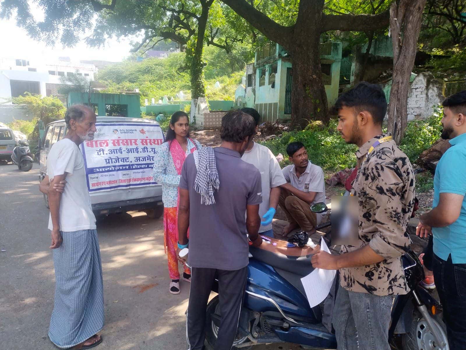 People around a scooter at the Shisha Khan camp, beside a van carrying the TI-IDU project's banner
