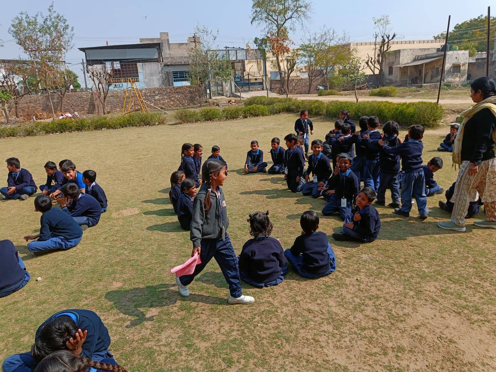 BSPS students sitting outdoors on the school field during a group activity, with a teacher standing nearby
